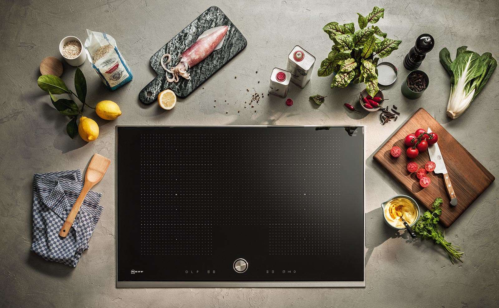 Flex-Induktionskochfeld von Neff mit schwarzer Glasoberfläche und digitaler Steuerung, umgeben von frischen Zutaten und Küchenutensilien auf grauer Arbeitsfläche.