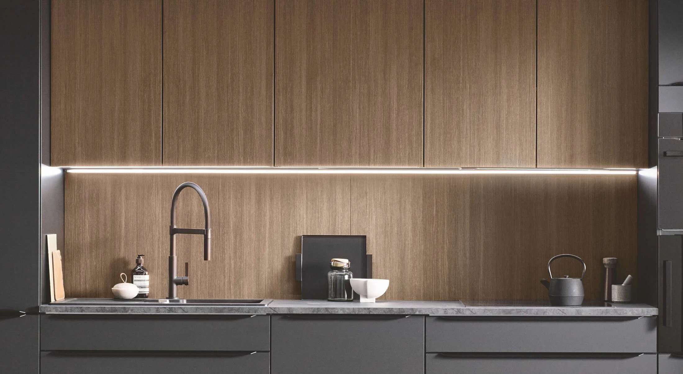 Dunkle Küchenzeile mit Holzrückwand, durchgehendem Lichtband und schwarzer Armatur in einer modernen Burger Küche.